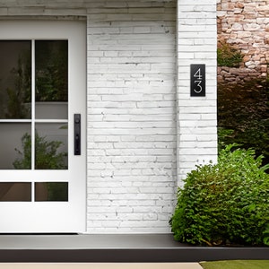 The house number sign is shown from a distance on a house made of brick. The numbers on the plaque are clearly visible and the proportions look appropriate for the home. Shrubs and a grassy lawn make this a beautiful scene around the sign.