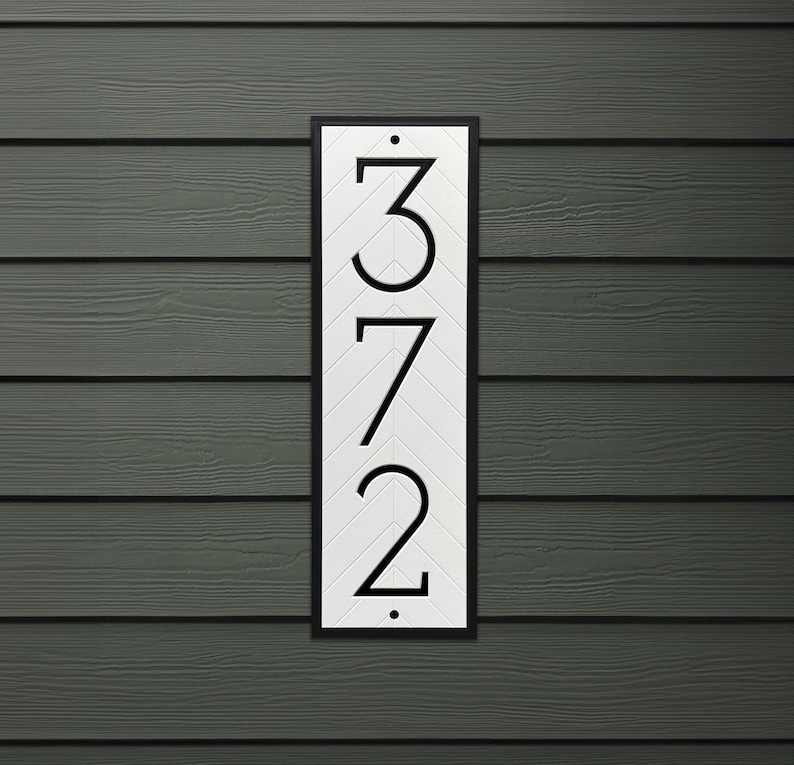 Vertical address plaque is mounted to vinyl siding. The house number sign is white with a black border and black modern numbers that are recessed into address sign. A subtle, white herringbone pattern fills the white background inside the frame.