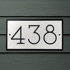 Horizontally oriented, rectangular address plaque is mounted to vinyl siding. The sign is white with a black border and black modern numbers recessed into the white sign. A subtle, white chevron pattern fills the white background inside the frame.