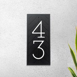 A rectangular address plaque is mounted vertically to a white wall. The plaque is black with white, modern house numbers recessed into plaque. A black, recessed chevron pattern is around the numbers and fills the rectangular shape of the sign.