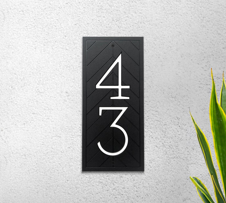 A rectangular address plaque is mounted vertically to a white wall. The plaque is black with white, modern house numbers recessed into plaque. A black, recessed chevron pattern is around the numbers and fills the rectangular shape of the sign.