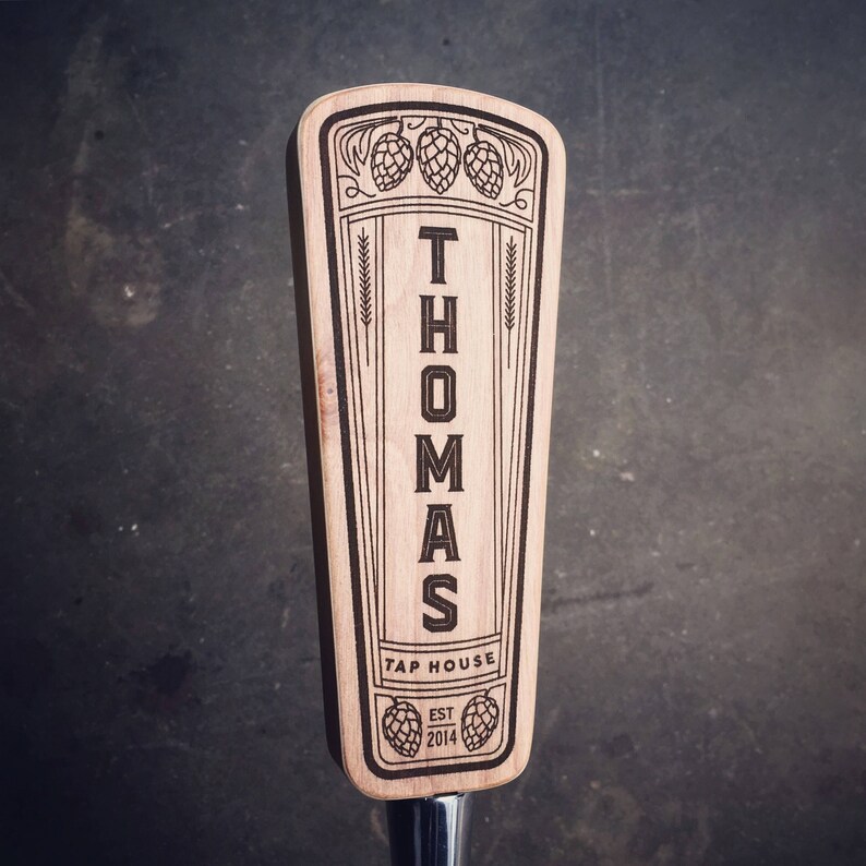 May include: A wooden beer tap handle with the name "Thomas Tap House" and the year "2014" engraved on it. The handle is decorated with a stylized design of hops and wheat.