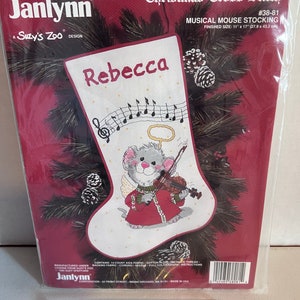 May include: A red and white cross stitch pattern for a Christmas stocking featuring a musical mouse playing a violin. The stocking is 28 cm by 43 cm and includes the name "Rebecca".