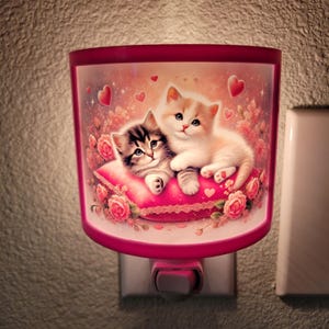 May include: A pink nightlight with a white plug-in base. The nightlight features a picture of two kittens cuddling on a pink pillow with hearts and flowers.