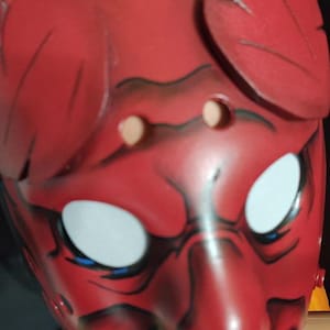 May include: A red Oni mask with white eyes, blue accents, and a menacing expression. The mask features black detailing and two leaf-like protrusions on top. The mask has two eye holes and a mouth with visible teeth.
