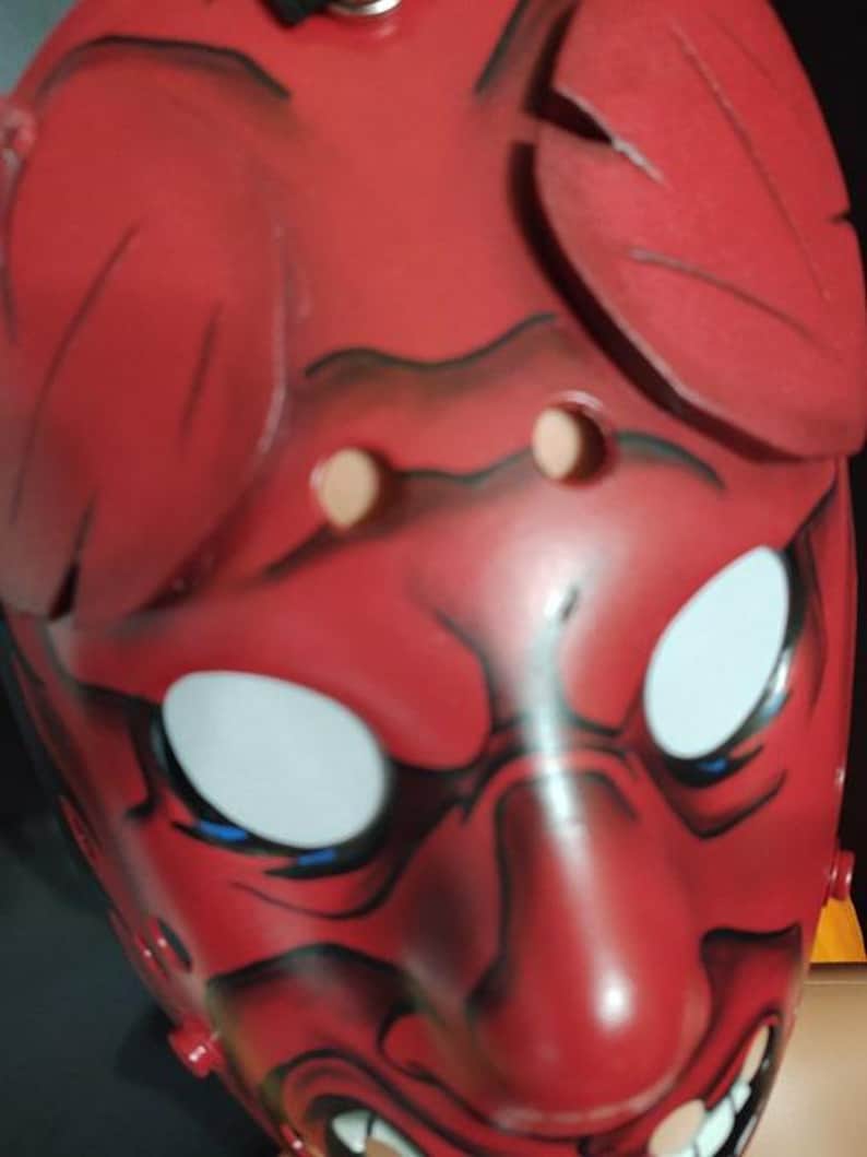 May include: A red Oni mask with white eyes, blue accents, and a menacing expression. The mask features black detailing and two leaf-like protrusions on top. The mask has two eye holes and a mouth with visible teeth.