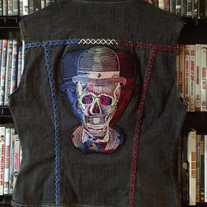 May include: Dark denim vest with a skull design on the back. The skull is embroidered with red, white, and blue thread, and is wearing a top hat. The vest has blue and red stitching details.