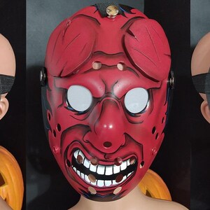 May include: A red, stylized mask with white eyes and a menacing grin. The mask features black accents and a strap for wearing. The design includes decorative elements on the forehead and cheeks, creating a striking visual.
