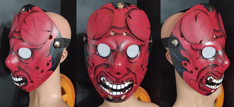 May include: A red, stylized mask with white eyes and a menacing grin. The mask features black accents and a strap for wearing. The design includes decorative elements on the forehead and cheeks, creating a striking visual.