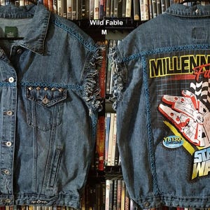 May include: Blue denim jacket with a distressed look, featuring a studded front and frayed edges. The back displays a colorful graphic of the Millennium Falcon from Star Wars, with the text "MILLENNIUM FALCON" and "STAR WARS".
