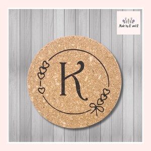 May include: Circular cork coaster featuring the letter "K" in black, encircled by a black frame adorned with heart motifs. The coaster is branded with the text "Made by R and R" in the top right corner.