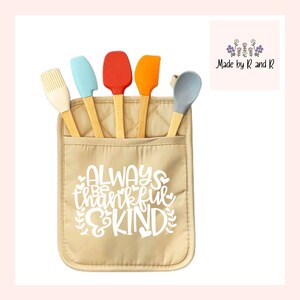 May include: Beige quilted hot pad with a pocket for utensils. The pocket contains five silicone utensils with wooden handles: a red spatula, an orange spatula, a blue spatula, a light blue spoon, and a white whisk. The hot pad has the text "Always be thankful & kind" printed on it in white.