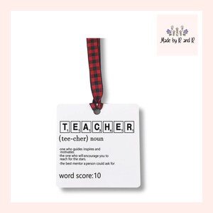 May include: A white square ornament with the word "TEACHER" spelled out in Scrabble tiles. The ornament includes a definition and a red and black plaid ribbon for hanging. The text reads "word score: 10".