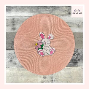 Könnte beinhalten: Ein rosa gewebter Platzdeckchen mit einem weißen Hasen, der eine rosa Blume hält, die darauf gestickt ist.
