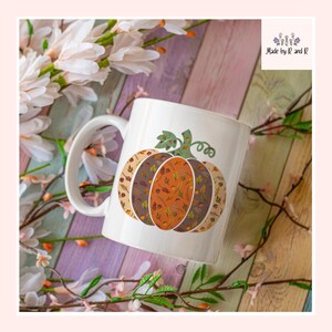 Puede incluir: Taza de cerámica blanca con un diseño de calabaza en tonos marrón, beige y naranja, decorada con hojas. La taza tiene un asa curva y el texto "Made by R and R". La taza está rodeada de flores y ramas blancas.