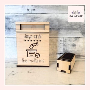 Può includere: Una scatola di legno per il conto alla rovescia con la scritta "days until the midterms" e una grafica di voto. Accanto c'è una piccola scatola rettangolare di legno. Lo sfondo è un motivo a venature di legno chiaro.