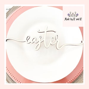 May include: A white wooden "Easter" sign with a cursive font on a pink plate. The sign is ideal for adding a festive touch to your Easter table.