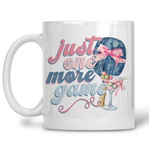 May include: White ceramic mug with the text "just one more game" in pink and blue script. The mug features a watercolor illustration of a blue object with a pink bow and a martini glass with colorful spheres.
