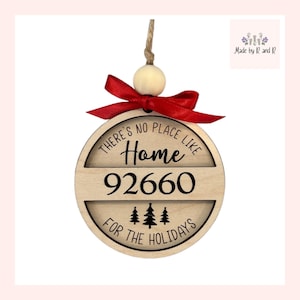 Puede incluir: Un adorno navideño redondo de madera con una cinta roja. El adorno está decorado con el texto "There's no place like home 92660 for the holidays".