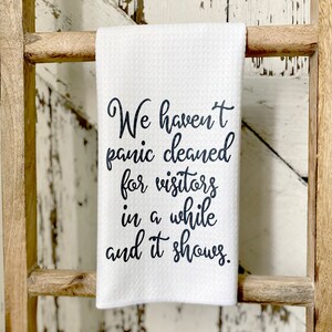 May include: White kitchen towel with black text that reads "We haven't panic cleaned for visitors in a while and it shows."