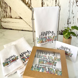 May include: A gift set of four white kitchen towels with the text "Happy Birthday" printed in colourful, stylised lettering. The towels are packaged in a brown box with a clear window.
