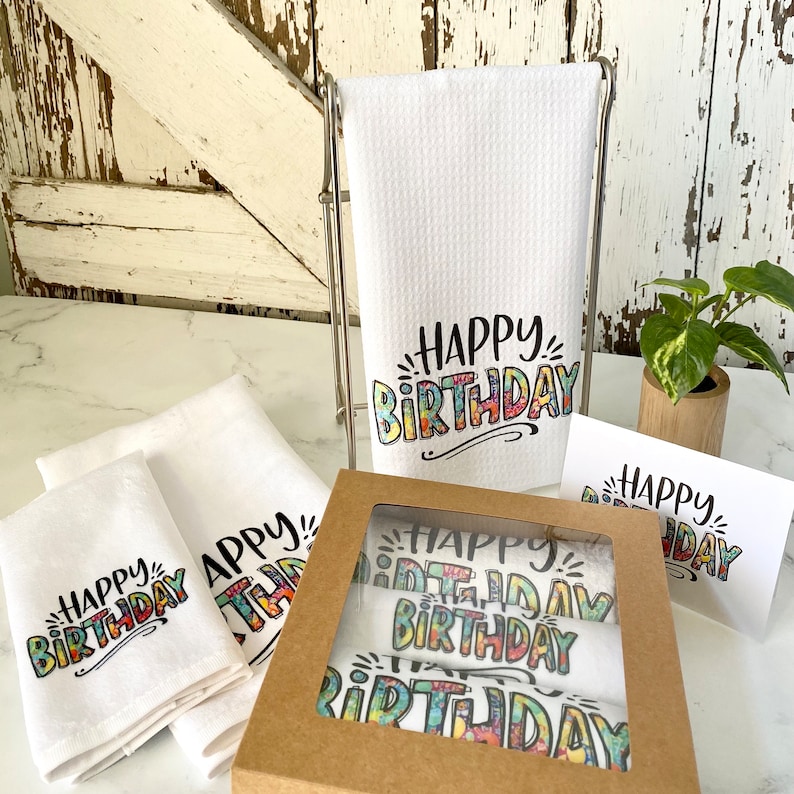May include: A gift set of four white kitchen towels with the text "Happy Birthday" printed in colourful, stylised lettering. The towels are packaged in a brown box with a clear window.