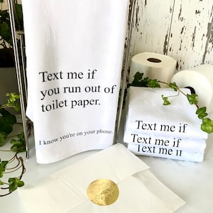 May include: White kitchen towels with the text "Text me if you run out of toilet paper. I know you're on your phone." printed on them. The towels are folded and stacked on a white surface.