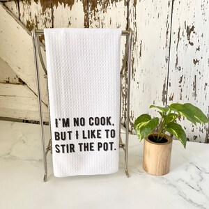 May include: White kitchen towel with black text that reads "I'm no cook, but I like to stir the pot." The towel is hanging on a silver towel rack. A small potted plant is on a white countertop.