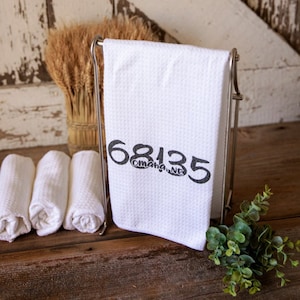 May include: White kitchen towel with black text that reads "68135 Omaha, NE" hanging on a metal towel rack.