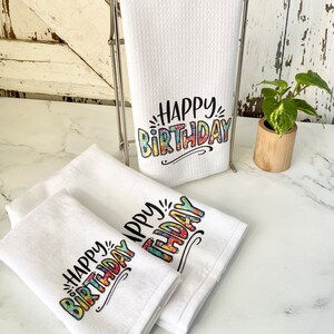 May include: Three white kitchen towels with the text "Happy Birthday" printed in colourful, patterned letters. The towels are folded and stacked on a white countertop.