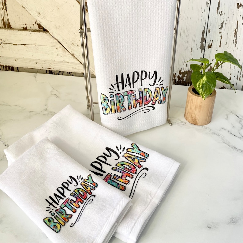 May include: Three white kitchen towels with the text "Happy Birthday" printed in colourful, patterned letters. The towels are folded and stacked on a white countertop.