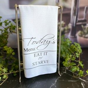 May include: White kitchen towel with the text "Today's Menu: Eat It or Starve" in black script. The towel is hanging on a silver metal rack, with green plants in the background. The towel has a textured surface.