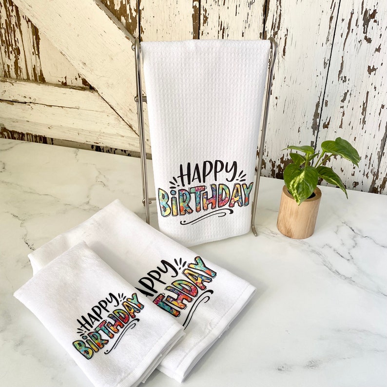 May include: Three white kitchen towels with the text "Happy Birthday" printed in colourful letters. The towels are folded and displayed on a white countertop.