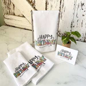 Three white kitchen towels with the words 'Happy Birthday' printed on them in colorful letters. The towels are folded and stacked on a white surface. A card with the same text is also visible in the image.