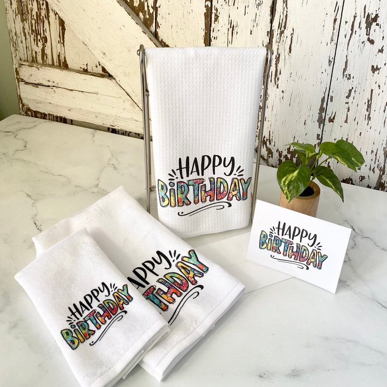 Three white kitchen towels with the words 'Happy Birthday' printed on them in colorful letters. The towels are folded and stacked on a white surface. A card with the same text is also visible in the image.