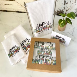 May include: A set of four white kitchen towels with the phrase "Happy Birthday" printed in colourful, rainbow-like lettering. The towels are packaged in a brown box with a clear window.