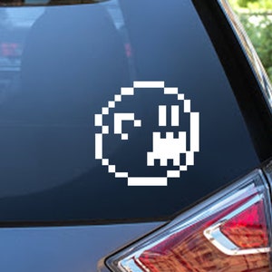 Puede incluir: Una pegatina de cara de fantasma pixelada blanca en una ventana de coche gris oscuro.