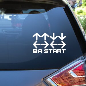 May include: A black car with a white sticker on the back window. The sticker has arrows pointing in different directions and the text "BA START".