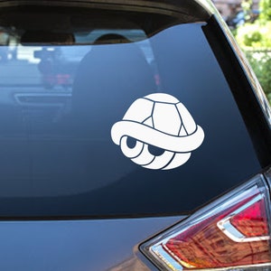 Op de afbeelding: Een witte sticker van een videogamepersonage, een schildpadenschild, op de achterruit van een grijze auto.
