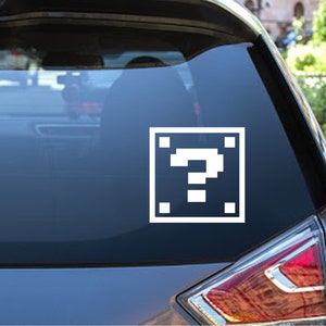 Puede incluir: Una pegatina blanca de un signo de interrogación pixelado dentro de un cuadrado en la ventana de un coche.