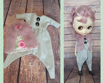 May include: A doll wearing a striped jumpsuit and a pink faux fur vest. The doll has pink hair and is wearing glasses.