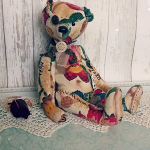 May include: A handmade patchwork teddy bear with a floral pattern in shades of red, green, and yellow. The bear has a button eye and a bow tie. The bear is sitting on a white lace doily.