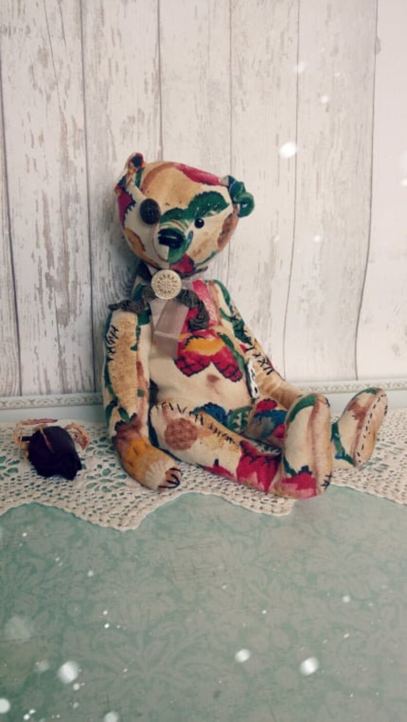 May include: A handmade patchwork teddy bear with a floral pattern in shades of red, green, and yellow. The bear has a button eye and a bow tie. The bear is sitting on a white lace doily.