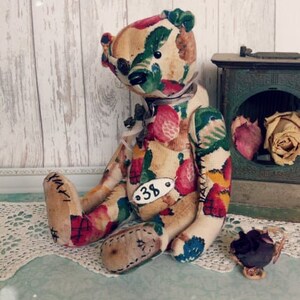 May include: A handmade teddy bear with a floral fabric pattern. The bear is sitting on a white lace doily and has a tag with the number "38" on its chest.
