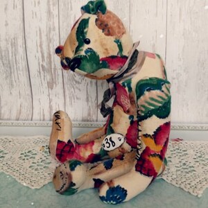May include: A handmade patchwork teddy bear with a floral print in shades of red, green, yellow, and blue. The bear is sitting on a white lace doily and has a tag with the number "38" on its leg.