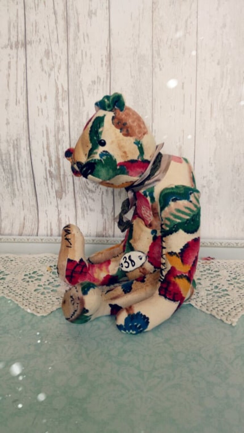 May include: A handmade patchwork teddy bear with a floral print in shades of red, green, yellow, and blue. The bear is sitting on a white lace doily and has a tag with the number "38" on its leg.