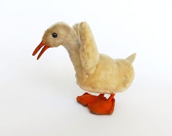Könnte beinhalten: Ein Vintage-Plüschtier in Form einer Ente. Es hat einen cremefarbenen Körper, einen orangefarbenen Schnabel und orangefarbene Schwimmfüße. Die Ente hat einen langen Hals und steht auf einer weißen Oberfläche. Das Spielzeug hat ein klassisches, nostalgisches Aussehen.