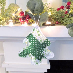 May include: A handmade Christmas tree ornament. The tree is made of green and white patterned fabric, with a patchwork design. The ornament is hanging from a white string. The background includes greenery, red berries, and lights.