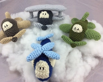 May include: Four crocheted airplane toys in different colors: brown, gray, green, and blue. The toys are sitting on a white fluffy surface.