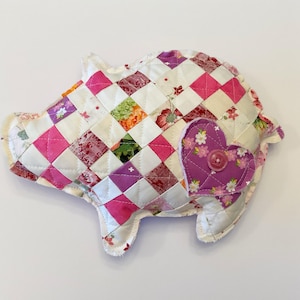 May include: A handmade, quilted pig-shaped pillow. The pig is constructed from a patchwork of pink, white, and floral patterned squares. A purple heart with a button is sewn on the side. The pillow is a charming decorative item.